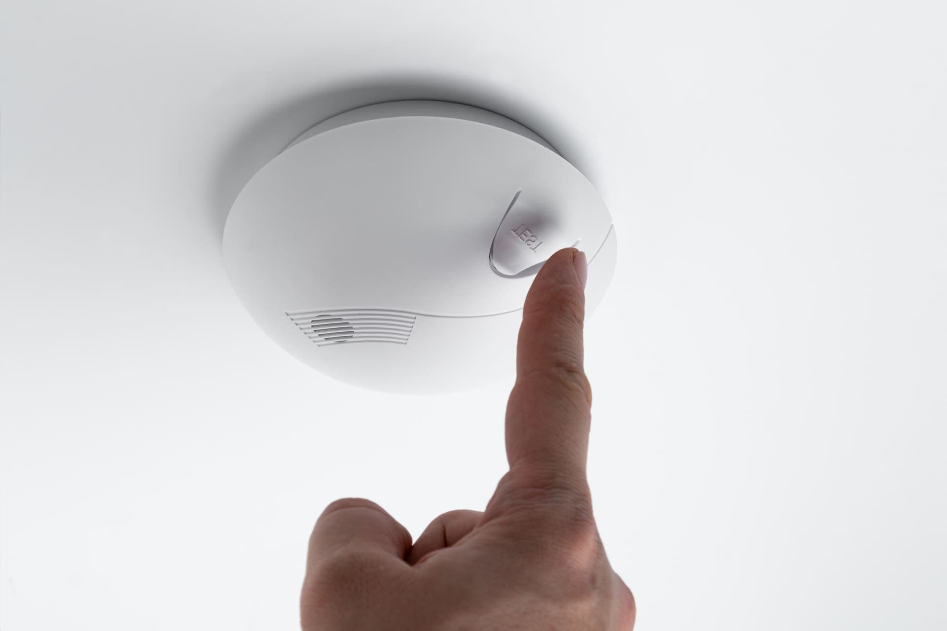 A finger pressing the test button on a ceiling smoke alarm